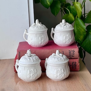 Puede incluir: Cuatro tarros blancos de cerámica con tapa y asas, con diseños florales en relieve. Cada tarro tiene un adorno en forma de tulipán en la tapa. Los tarros están colocados encima de dos libros rojos.