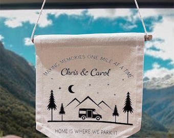 May include: A beige canvas banner with the text "MAKING MEMORIES ONE MILE AT A TIME" and "Chris & Carol". Below the text is a graphic of a camper van, mountains, trees, and a crescent moon. The banner reads "HOME IS WHERE WE PARK IT".