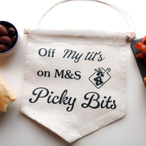 Può includere: Un banner di tela color crema con il testo "Off my tit's on M&S Picky Bits" e una grafica di tagliere. L'immagine include anche una baguette, olive in una ciotola blu e involtini di salsiccia.