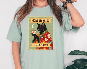 May include: A light green t-shirt featuring a vintage-style graphic. The graphic depicts a black cat in a kimono holding a cup of coffee, with the text "MORE ESPRESSO" above and "LESS DEPRESSO" below.
