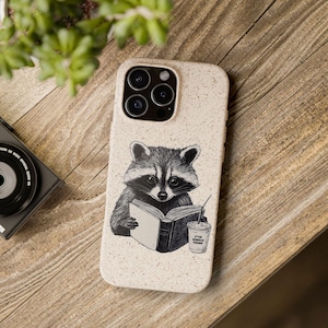 May include: A beige phone case featuring a black and white illustration of a raccoon reading a book. The raccoon is holding a book and a cup of coffee with the text "F*ck Donald Trump". The phone case is on a wooden surface.