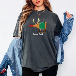 May include: Dark gray t-shirt featuring a cartoon illustration of a bottle with antlers and a green label, resembling a reindeer. The text "Brew Dolf" is printed below the graphic in white. The shirt is a comfortable, casual style.