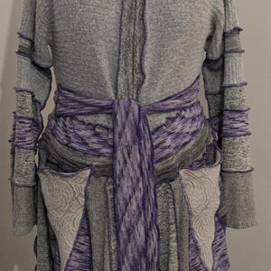 May include: A gray hooded cardigan with purple accents, displayed on a mannequin. The long-sleeved sweater features a tie belt, decorative pockets, and contrasting purple trim. The design includes a hood and a flowing, asymmetrical hemline.
