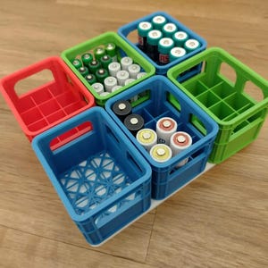 Puede incluir: Una colección de pequeñas cajas de plástico de colores, cada una llena de pilas. Las cajas son rojas, verdes y azules, y las pilas son de varios tamaños y colores. Las cajas están dispuestas sobre una superficie de madera.