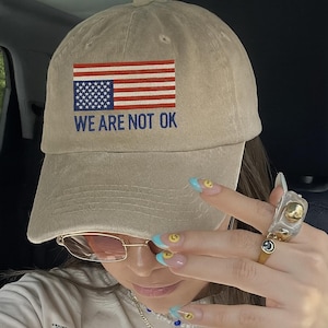 May include: A beige baseball cap with an American flag graphic and the text "WE ARE NOT OK". The image also shows a white t-shirt with a graphic print and a necklace.