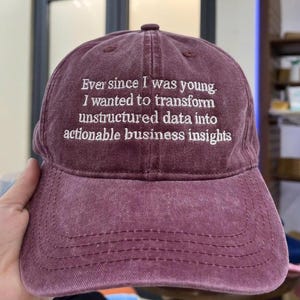 May include: A maroon baseball cap with white embroidered text. The text reads, "Ever since I was young, I wanted to transform unstructured data into actionable business insights." The cap has a curved peak and a button on top.