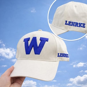 May include: A cream-colored baseball cap with a large blue embroidered "W" on the front. The name "LEHRKE" is embroidered in blue on the side of the cap. The cap is set against a blue sky background.
