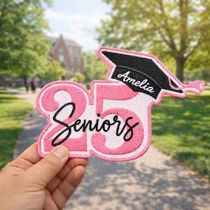 Op de afbeelding: Een roze en witte geborduurde patch met de tekst "25 Seniors" en een afstudeerhoed met de naam "Amelia". De patch wordt in een hand gehouden tegen een vage buitenachtergrond.