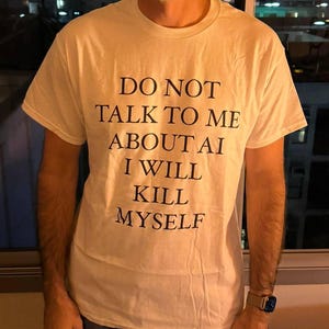 Könnte beinhalten: Ein weißes T-Shirt mit dem Text "DO NOT TALK TO ME ABOUT AI I WILL KILL MYSELF" in Schwarz. Das Shirt wird mit einer blauen Jeans getragen.