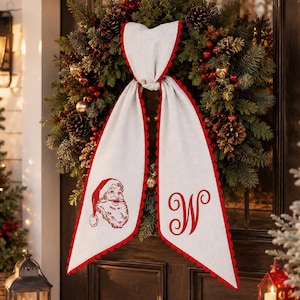 May include: A Christmas wreath made of green pine branches, pinecones, and red berries, featuring a white fabric ribbon with red scalloped edges. The ribbon displays a red illustration of Santa Claus on one side and a red letter W on the other.