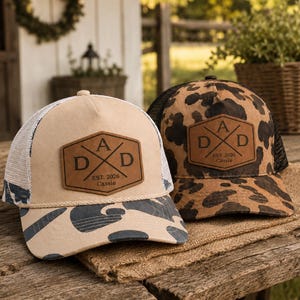Puede incluir: Dos gorras de camionero con parches de cuero. Una gorra es de color beige con una parte trasera de malla blanca y una visera de camuflaje. La otra gorra tiene un estampado de animales marrón y negro con una parte trasera de malla negra. Ambas gorras tienen el texto "A D D" y "EST. 2026 Cassie".