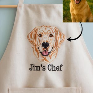 以下が含まれることがあります： ゴールデンレトリバーの顔が刺繍されたクリーム色のエプロン。「Jim's Chef」の文字が刺繍されています。犬の小さな画像が右上隅にあります。エプロンにはフロントポケットが付いています。