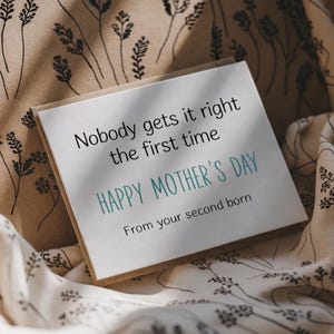 May include: A square greeting card with the text "Nobody gets it right the first time" and "HAPPY MOTHER'S DAY" in teal. The card is on a patterned fabric background, perfect for Mother's Day.