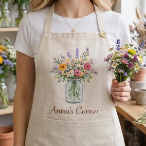 Peut inclure: Tablier beige avec une broderie florale dans un bocal en verre et le texte "Anna's Corner". Le tablier a deux poches et une fermeture à bouton en bois. Un bouquet de fleurs est tenu.