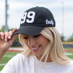 May include: Black baseball cap with the number "99" in white block letters and the name "Emily" embroidered in script. The cap is worn by a person wearing a white t-shirt.