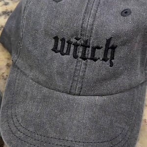 May include: A gray baseball cap with the word "witch" embroidered in black gothic lettering. The cap has a curved brim and a fabric strap closure. The hat is made of a soft, worn material.