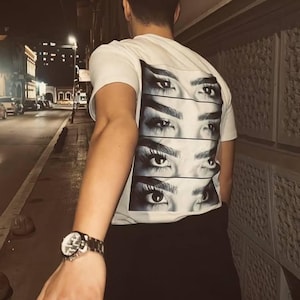 以下が含まれることがあります： 4組の目が描かれたグラフィックの白いTシャツ、黒いパンツとベージュのトラッカーハットを着用。夜の街を歩く人が、別の人の手を握っています。手首にはシルバーの腕時計があります。