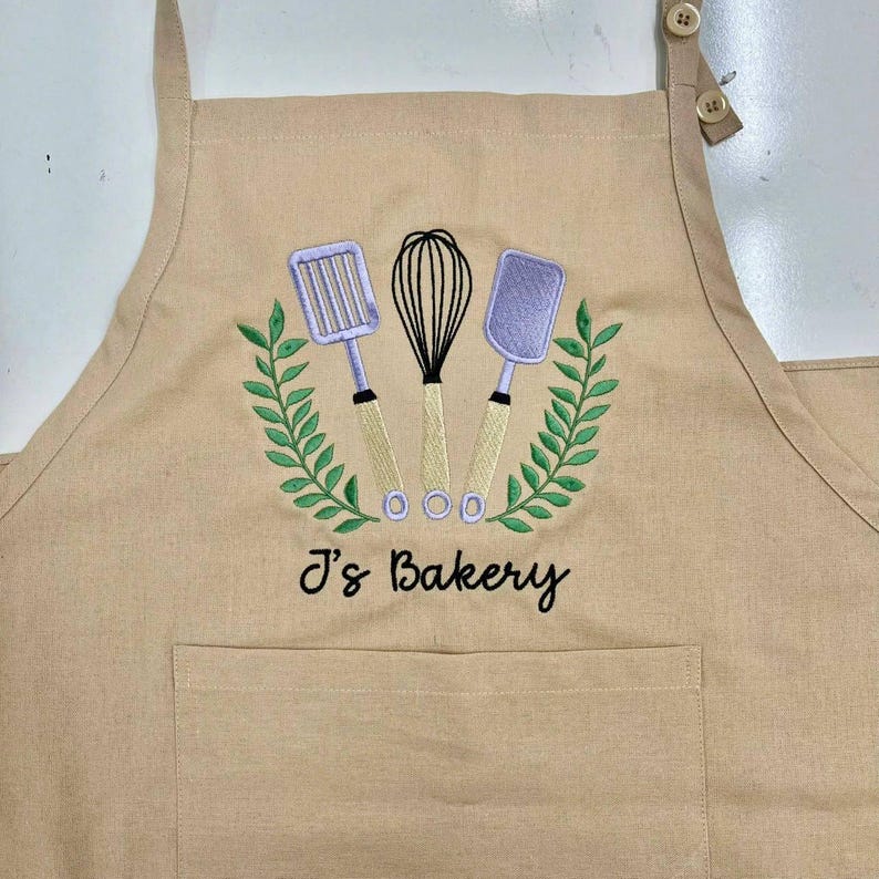 May include: Tan apron with an embroidered design featuring a whisk, spatula, and slotted turner, surrounded by a green wreath. The text "J's Bakery" is below the design. The apron has a front pocket and adjustable straps.