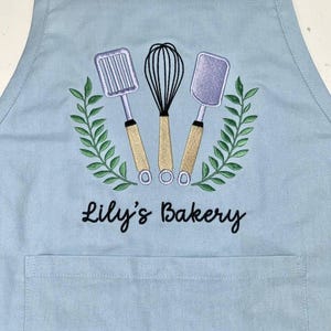 May include: A light blue apron with an embroidered design featuring a whisk, spatula, and slotted turner, surrounded by a green wreath. The text "Lily's Bakery" is embroidered below the design. The apron has a front pocket and adjustable neck strap.