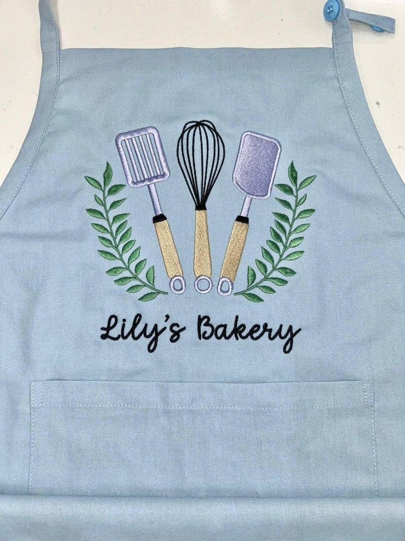 May include: A light blue apron with an embroidered design featuring a whisk, spatula, and slotted turner, surrounded by a green wreath. The text "Lily's Bakery" is embroidered below the design. The apron has a front pocket and adjustable neck strap.
