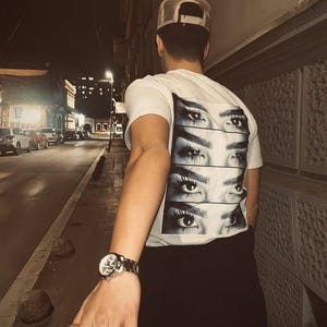Puede incluir: Una persona que lleva una camiseta blanca con un gr&aacute;fico de varios ojos en la espalda. La persona est&aacute; sosteniendo la mano de otra persona y caminando por una calle de noche.