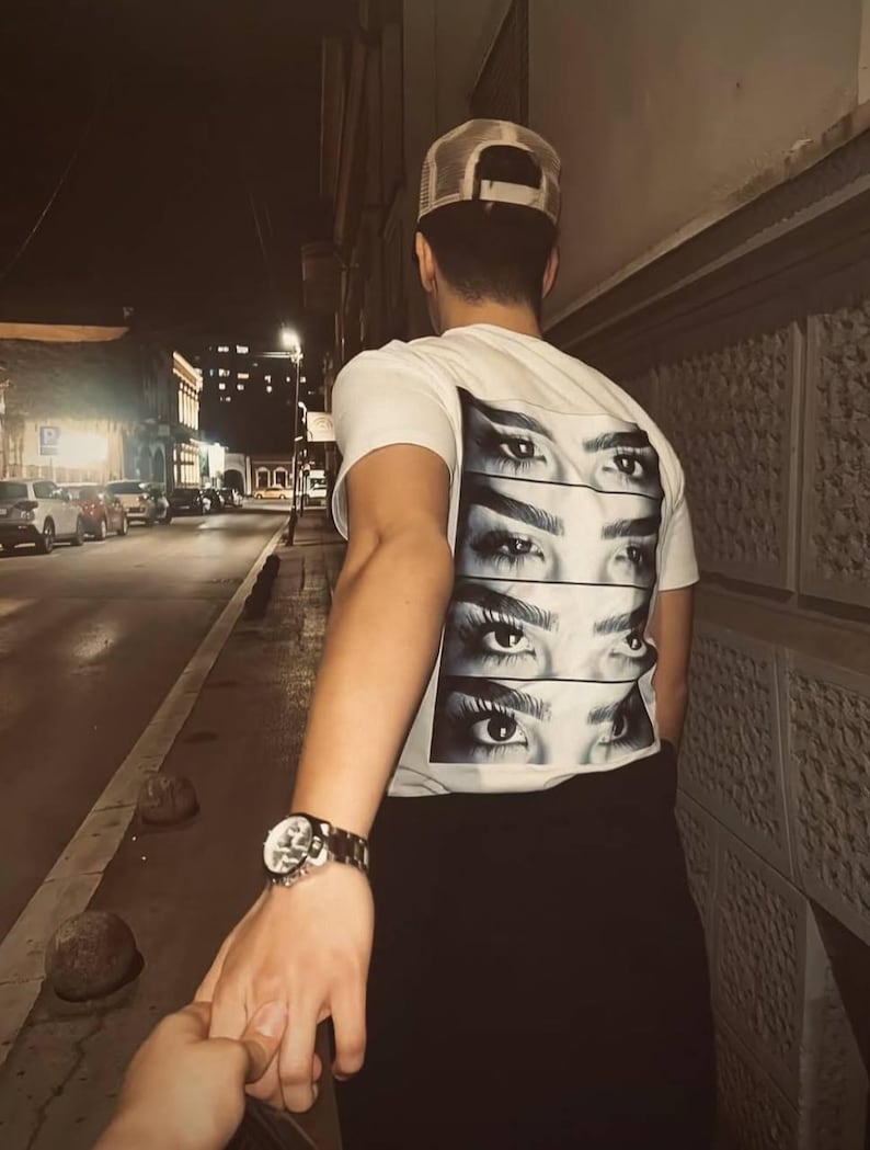 Puede incluir: Una persona que lleva una camiseta blanca con un gr&aacute;fico de varios ojos en la espalda. La persona est&aacute; sosteniendo la mano de otra persona y caminando por una calle de noche.