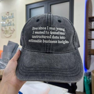 May include: A dark grey baseball cap with a curved peak. The cap has white embroidered text that reads, "Ever since I was young. I wanted to transform unstructured data into actionable business insights."