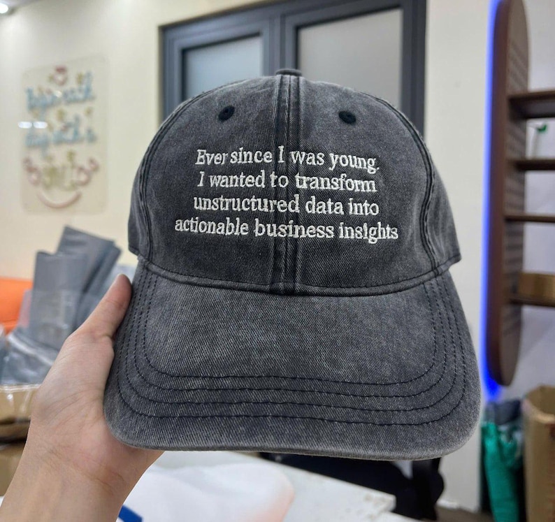 May include: A dark grey baseball cap with a curved peak. The cap has white embroidered text that reads, "Ever since I was young. I wanted to transform unstructured data into actionable business insights."