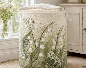 May include: A cylindrical quilted fabric laundry hamper featuring an off-white base with an intricate embroidered pattern of green lily of the valley stems, leaves, and white bell-shaped flowers, topped with a folded cream-colored knit blanket.