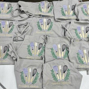 May include: A collection of gray aprons embroidered with kitchen utensils and names like "Zoe's Kitchen" and "Jennifer's Kitchen." Each apron features a whisk, spatula, and brush design within a green wreath.