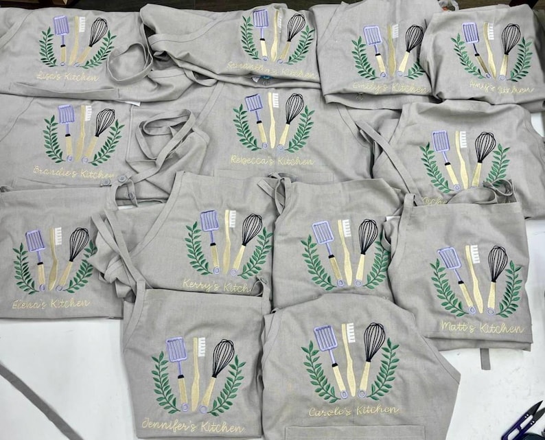 May include: A collection of gray aprons embroidered with kitchen utensils and names like "Zoe's Kitchen" and "Jennifer's Kitchen." Each apron features a whisk, spatula, and brush design within a green wreath.
