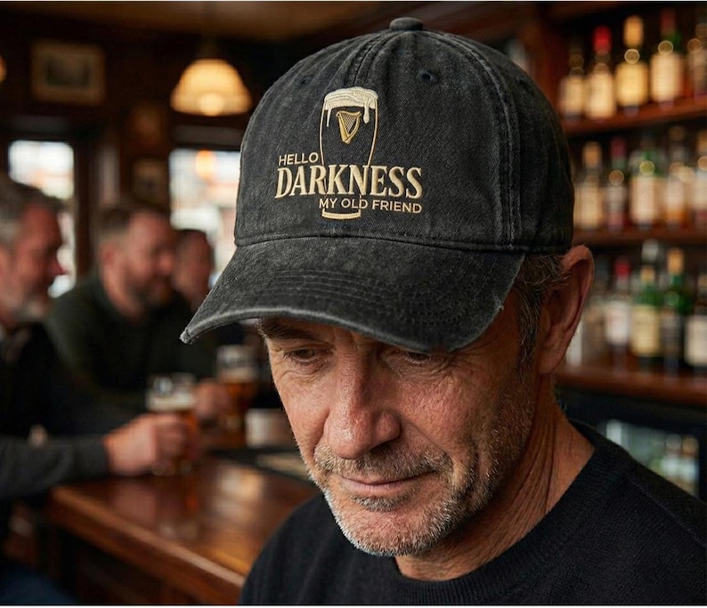 Może przedstawiać: Czarna czapka z daszkiem z napisem "Hello Darkness My Old Friend" w złotym kolorze. Czapka posiada logo Guinness. Zdjęcie zrobione w pubie.