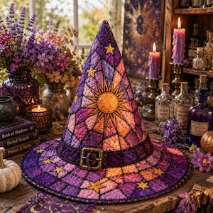 May include: A quilted witch hat featuring a stained-glass pattern in shades of purple, pink, and orange. The hat displays a central sun motif, scattered yellow stars, and a dark purple band with a metallic buckle at the base of the cone.
