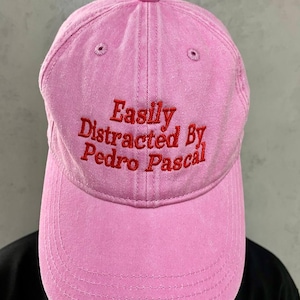 May include: A pink baseball cap featuring the embroidered phrase "Easily Distracted By Pedro Pascal" in red. The cap has a curved peak and a classic six-panel design. The hat is made of a soft, textured fabric.