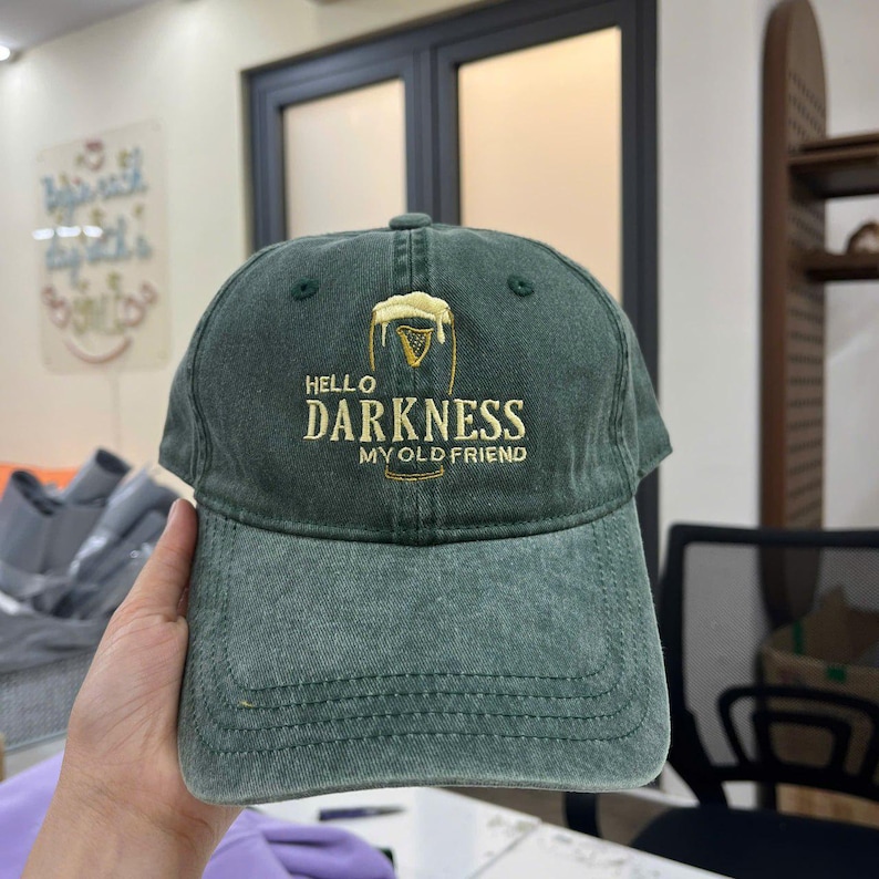 Może przedstawiać: Zielona czapka bejsbolowa z napisem "HELLO DARKNESS MY OLD FRIEND" w kolorze białym i złotym. Czapka posiada haftowany wz&oacute;r szklanki piwa. Kapelusz wykonany jest z miękkiego, zużytego materiału.