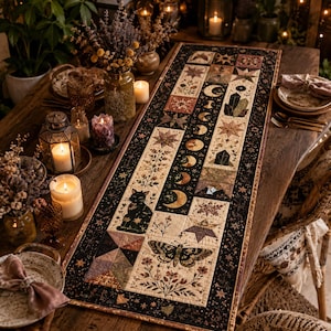May include: A rectangular fabric table runner featuring a patchwork design with a black border. The pattern includes illustrations of a black cat, a moth, floral motifs, stars, crystals, and a sequence of moon phases in shades of cream, brown, and black.