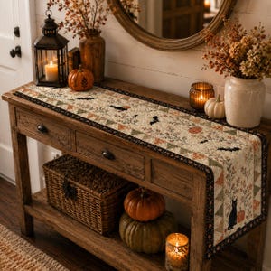 May include: A quilted table runner featuring a cream-colored fabric with a repeating pattern of black cats, bats, orange pumpkins, and autumn foliage. The runner is bordered by a dark fabric with a floral print and triangular geometric accents.