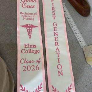 Może przedstawiać: Dwie białe satynowe szarfy absolwenckie z r&oacute;żowym wykończeniem i r&oacute;żowym haftem. Jedna szarfa zawiera napis Gloria Cruz, Bachelors of Science in Nursing, Elms College, Class of 2026 oraz symbol kaduceusza. Druga zawiera napis FIRST GENERATION. Widoczna linijka.