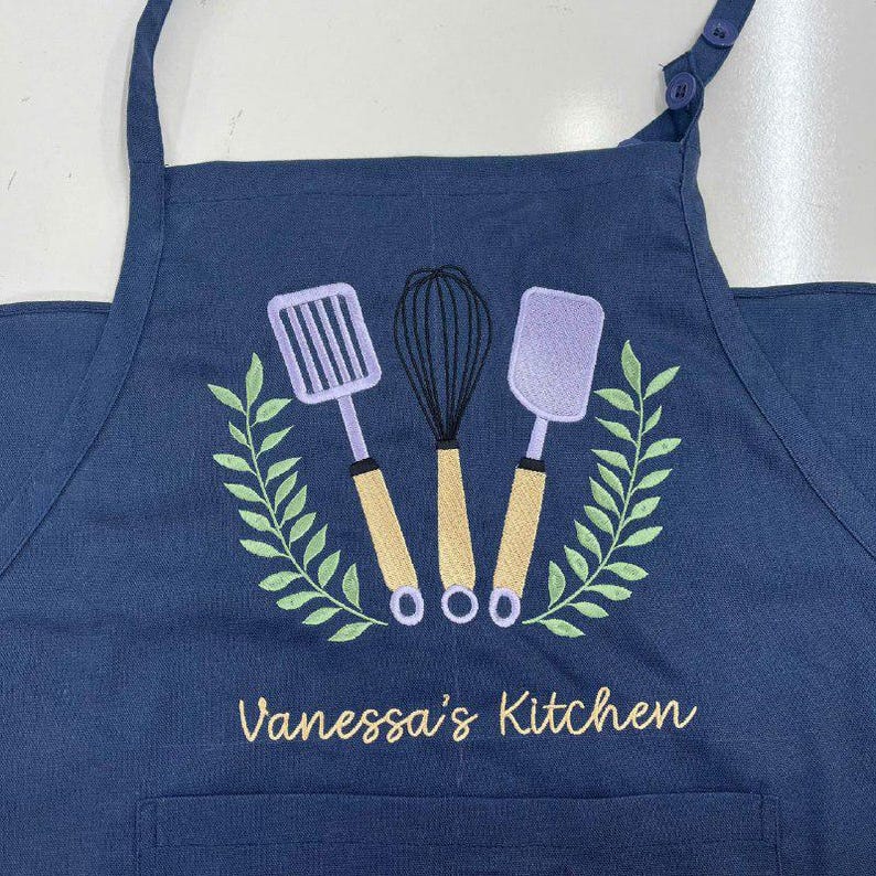 May include: A navy blue kitchen apron with an embroidered design featuring a whisk, spatula, and slotted turner. The design is surrounded by green leaves and the text "Vanessa's Kitchen" in gold thread.