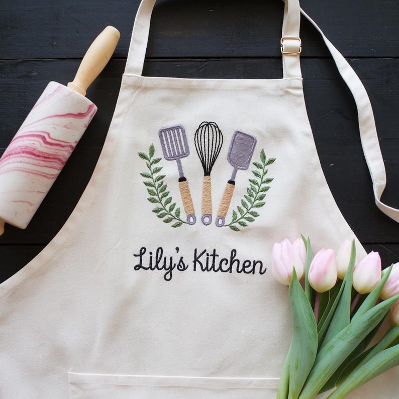 May include: Cream-colored apron with embroidered kitchen utensils and the text "Lily's Kitchen." A rolling pin with a pink and white marble design and a bouquet of pink tulips are also present.