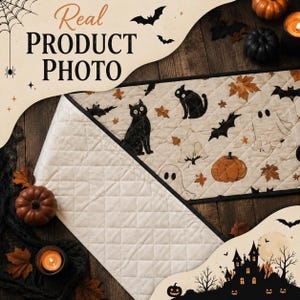 May include: A quilted table runner featuring a cream-colored fabric with a repeating pattern of black cats, white ghosts, orange pumpkins, bats, and brown autumn leaves. The runner has a diamond-stitched texture and a solid cream-colored underside.