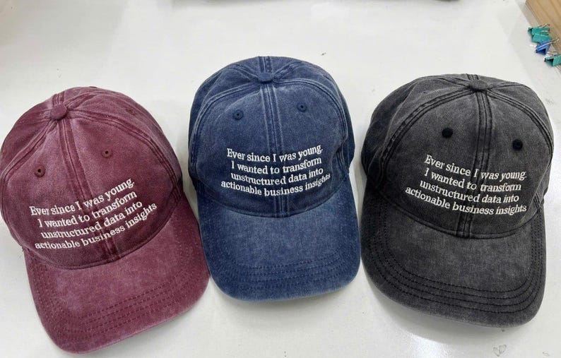 May include: Three baseball caps in burgundy, navy blue, and charcoal grey. Each cap has the embroidered text: "Ever since I was young I wanted to transform unstructured data into actionable business insights."