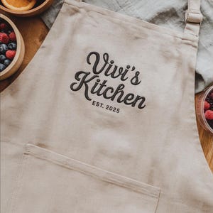 Op de afbeelding: Beige schort met een grote voorzak en verstelbare nekband. Het schort heeft de geborduurde tekst "Vivi's Kitchen" boven "EST. 2025" in zwart schrift. Het schort ligt op een houten oppervlak met kommen fruit.