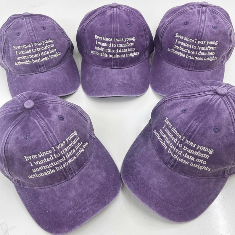 May include: Five purple baseball caps with white embroidered text: "Ever since I was young, I wanted to transform unstructured data into actionable business insights". The caps are made of a faded, textured fabric.