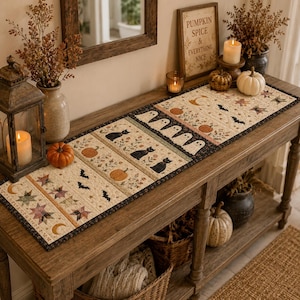 May include: A rectangular quilted table runner featuring a repeating pattern of black cats, white ghosts, orange pumpkins, black bats, and multicolored stars on a cream background with a dark border, displayed on a wooden console table.