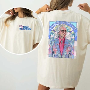 Pode incluir: Uma camiseta branca com um design de vitral azul e rosa nas costas. O design apresenta um homem com cabelo branco e uma camisa rosa, usando um terno azul estampado. O texto "Misbehavin'" está escrito em um arco azul acima do homem. A parte frontal da camiseta tem o texto "Baby Billy Bible Bonkers!" em azul e rosa.