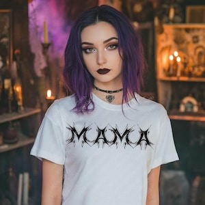 May include: A white t-shirt with the word "MAMA" in a black, gothic-style font. The shirt is worn by a person with purple hair and dark makeup. The background is dimly lit with candles and shelves.