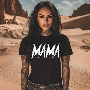 May include: Black t-shirt with the white word "MAMA" in a bold, jagged font. The shirt is short-sleeved and worn by a person with tattoos on their arms and neck. The background is a desert landscape.