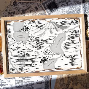 May include: A framed map of Icewind Dale, with a detailed black and white illustration of the region's geography. The map includes place names like Kelvin's Cairn, Bryn Shander, and Lac Dinneshere. The frame is made of light-coloured wood.