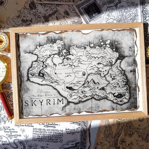 May include: A framed map of Skyrim from The Elder Scrolls V video game. The map is in black and white, with a distressed, aged appearance. A magnifying glass with a red handle and a compass are next to the frame.